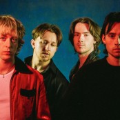 The four band members of Balancing Act stood in front of a blue background, with a red light shining towards them. Three are wearing black jackets, one is wearing a red one.